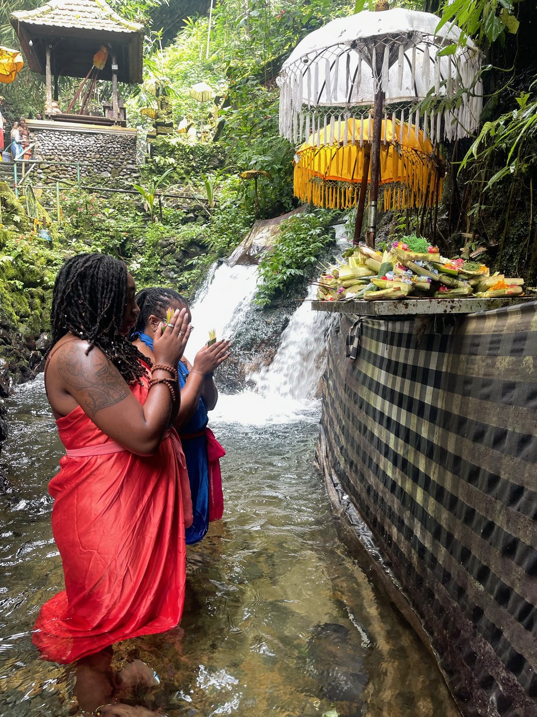 Ashleigh in a red sarong standing in a stream beneath a small waterfall shrine