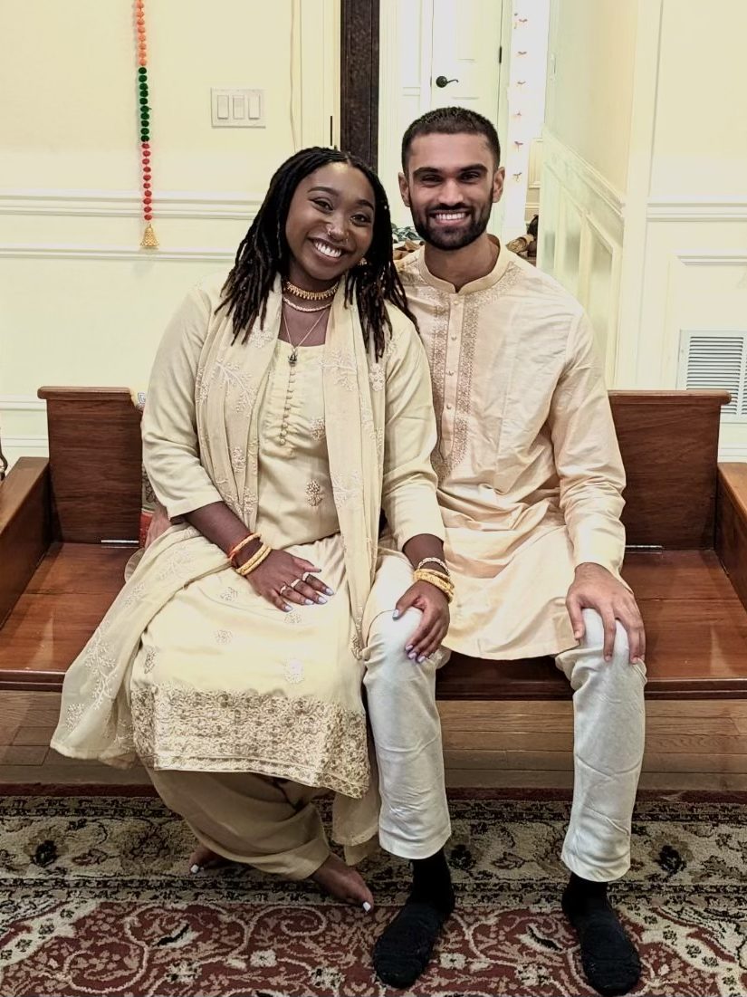 Ashleigh and Prithvi sitting together on a wooden swing, dressed in cream